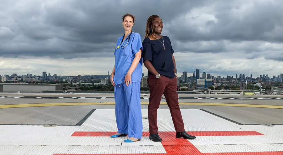 Picture of a doctor and a patient standing back to back on the helipad with dark grey sky and cityscape behind them