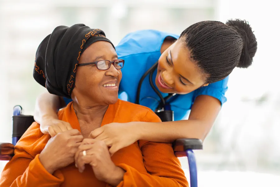 A patient in a wheelchair with a family member touching her shoulders behind her