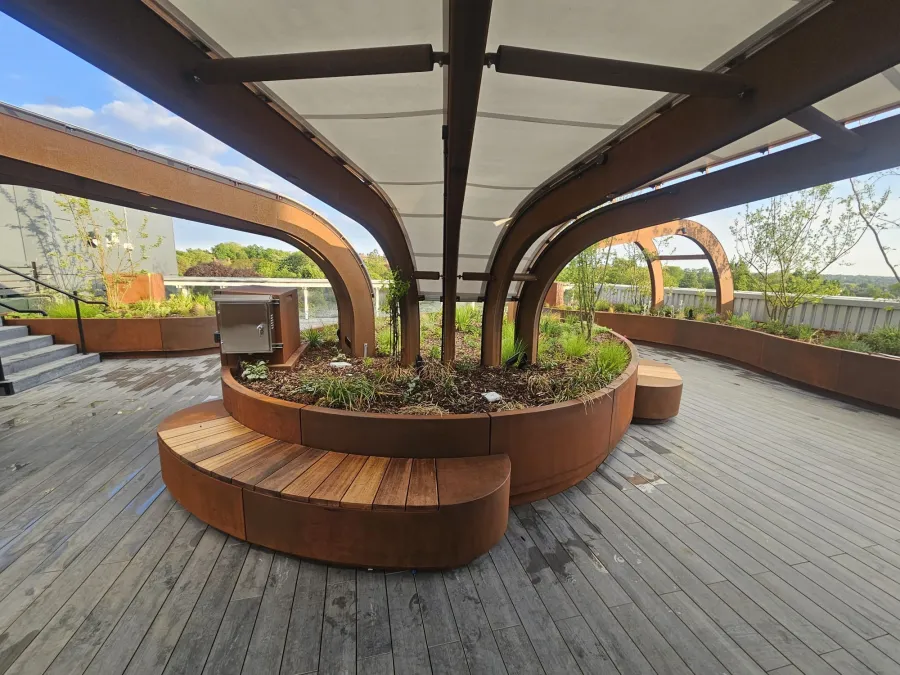 Curved wooden benches surround a central planter with metal arches and a canopy overhead on a rooftop deck.