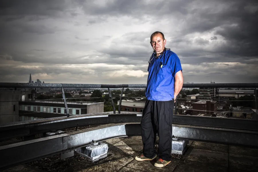 Dr Malcolm Tunnicliffe, Consultant in Emergency Medicine and Major Trauma at King’s College Hospital, standing on the helipad. He is wearing bright blue scrubs, which contrast strikingly to the stormy dark sky and clouds.