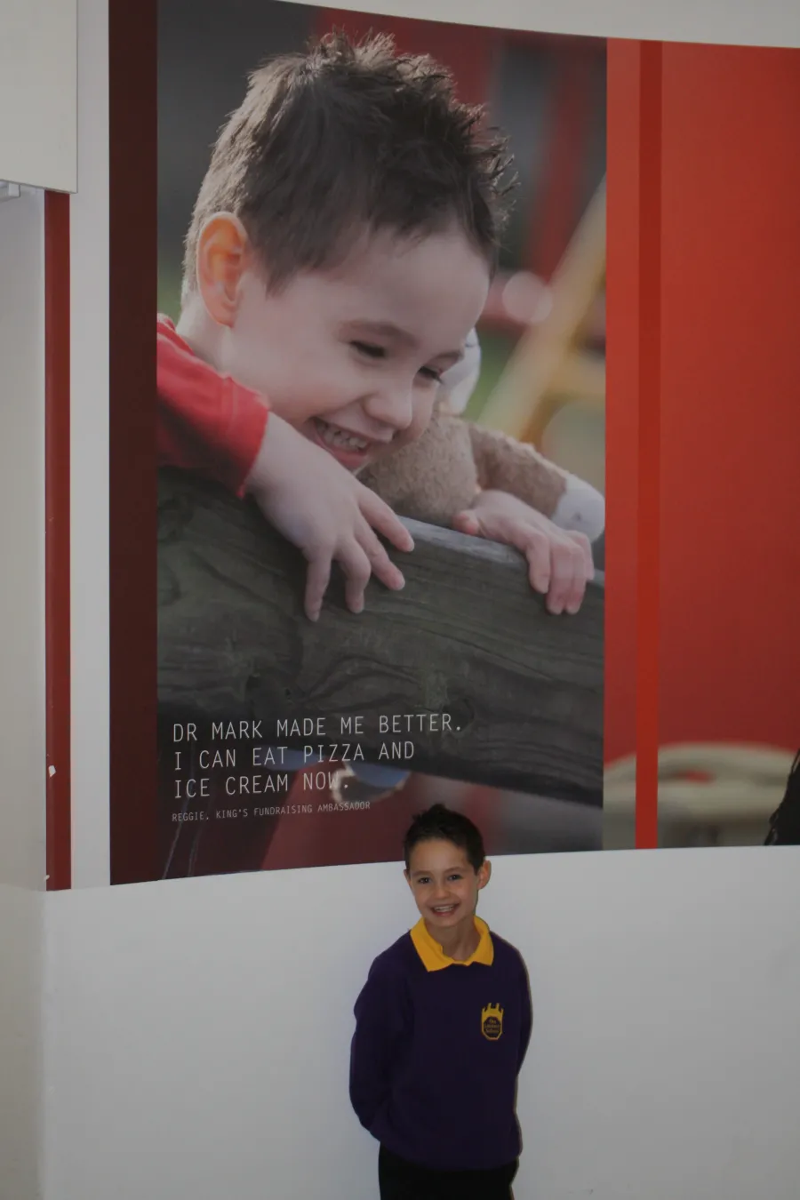 Reggie as a young child, standing in school uniform behind a large poster on the wall of him smiling, with a quote that reads: Dr Mark made me better. I can eat pizza and ice cream now.