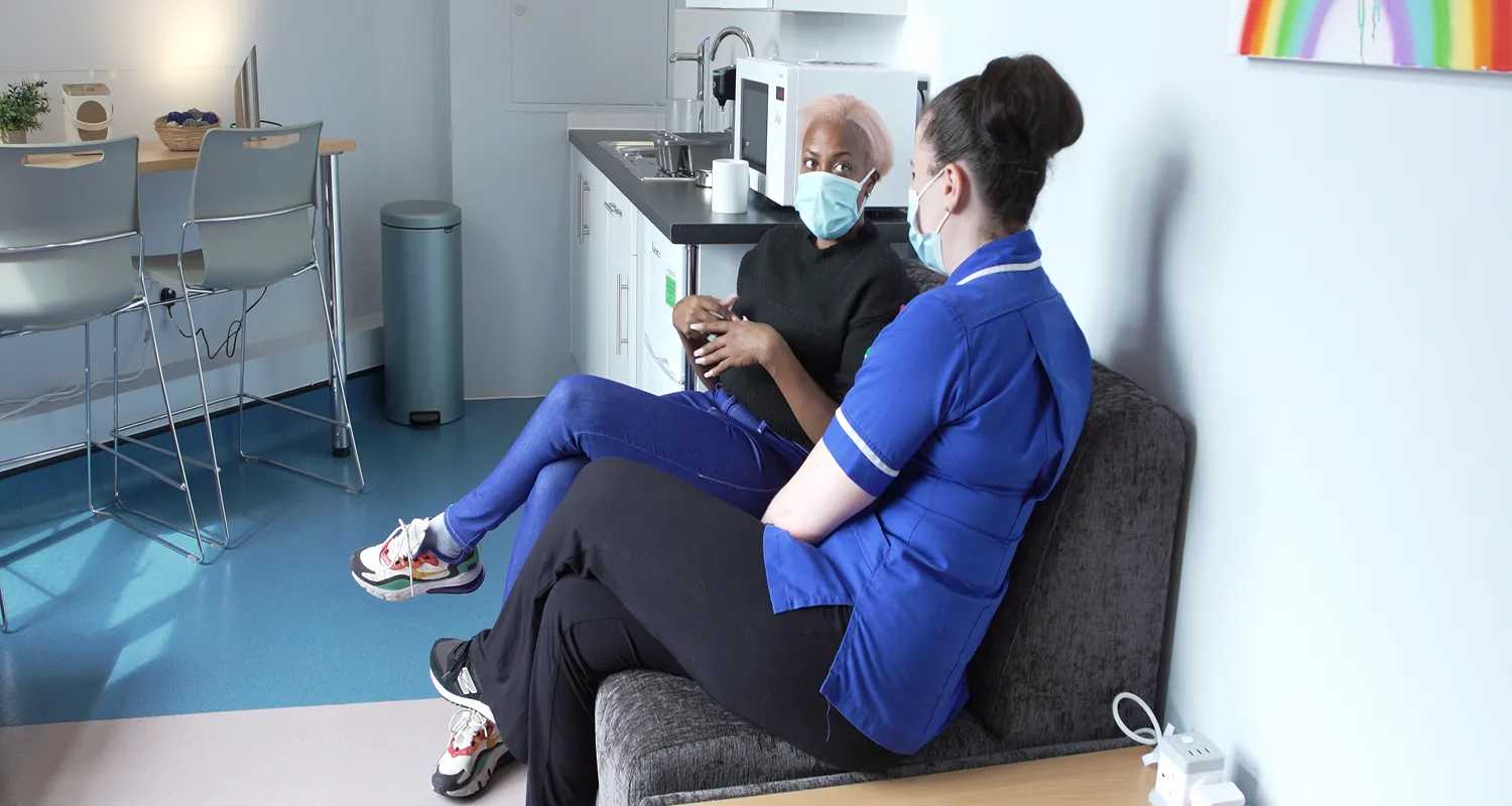 Aisha, sitting on a sofa with a nurse in the refurbished Toni & Guy parent's room. They are both wearing masks, and smiling in conversation. 