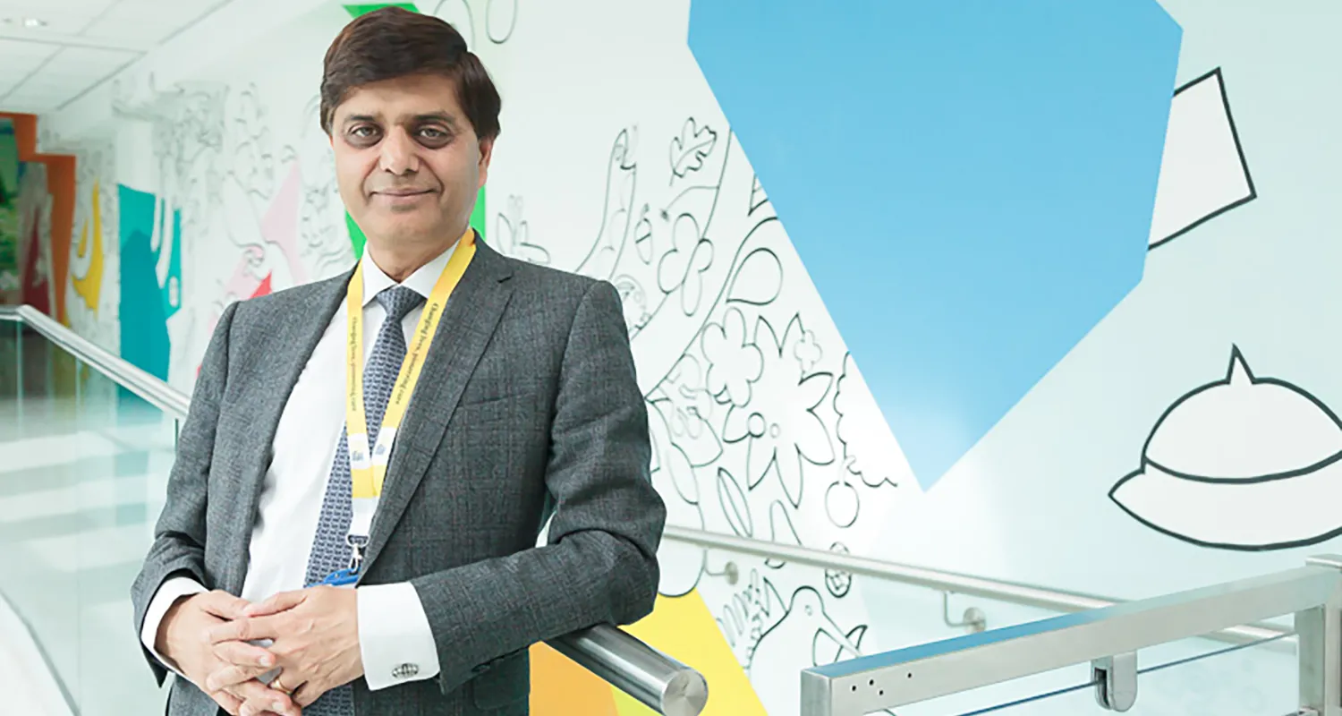Professor Anil Dhawan wearing a grey suit and blue tie, standing in a hospital corridor and smiling.