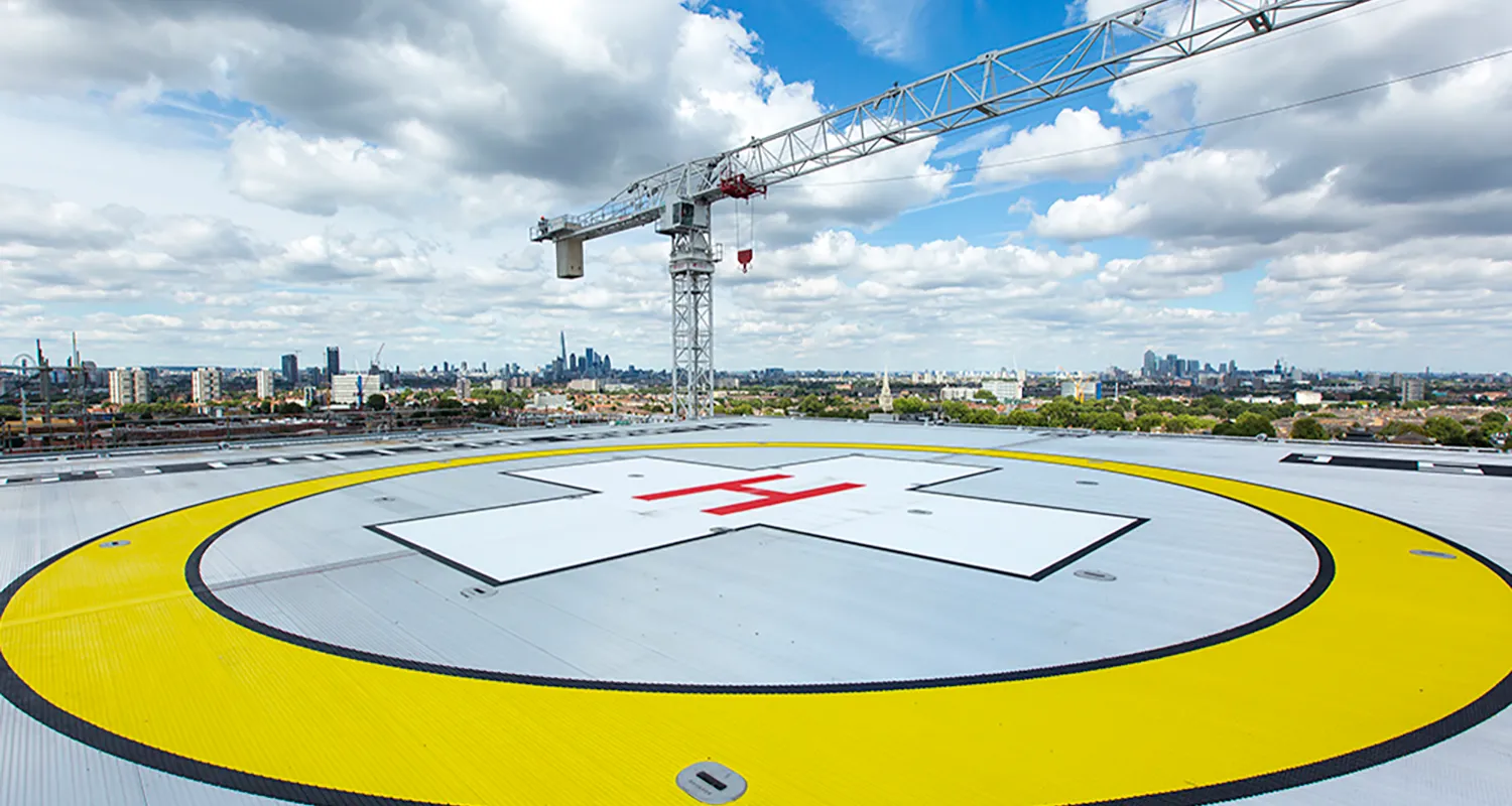 Birds-eye view of the helipad on a bright day with blue skies. The helipad has a bright yellow circle with a red cross in the middle to help air ambulances identify the landing spot. The London skyline is visible in the background.