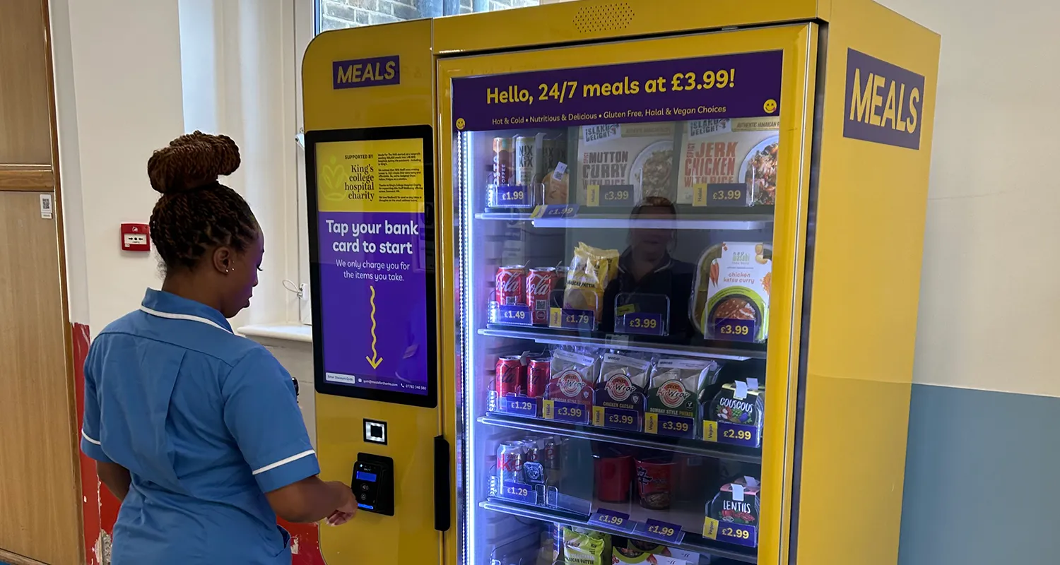 Female member of King's staff in blue overalls using the smart fridge in a hospital corridor. The smart fridge is bright yellow and has charity branding on it.