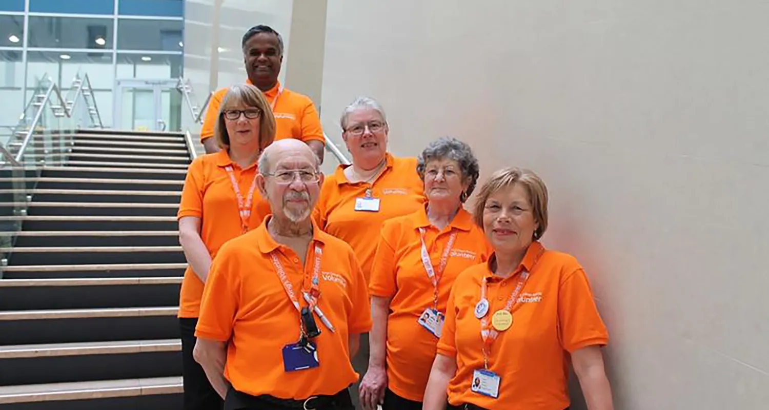 Six King's volunteers, wearing bright orange t-shirts, standing and smiling on the stairs of King's College Hospital