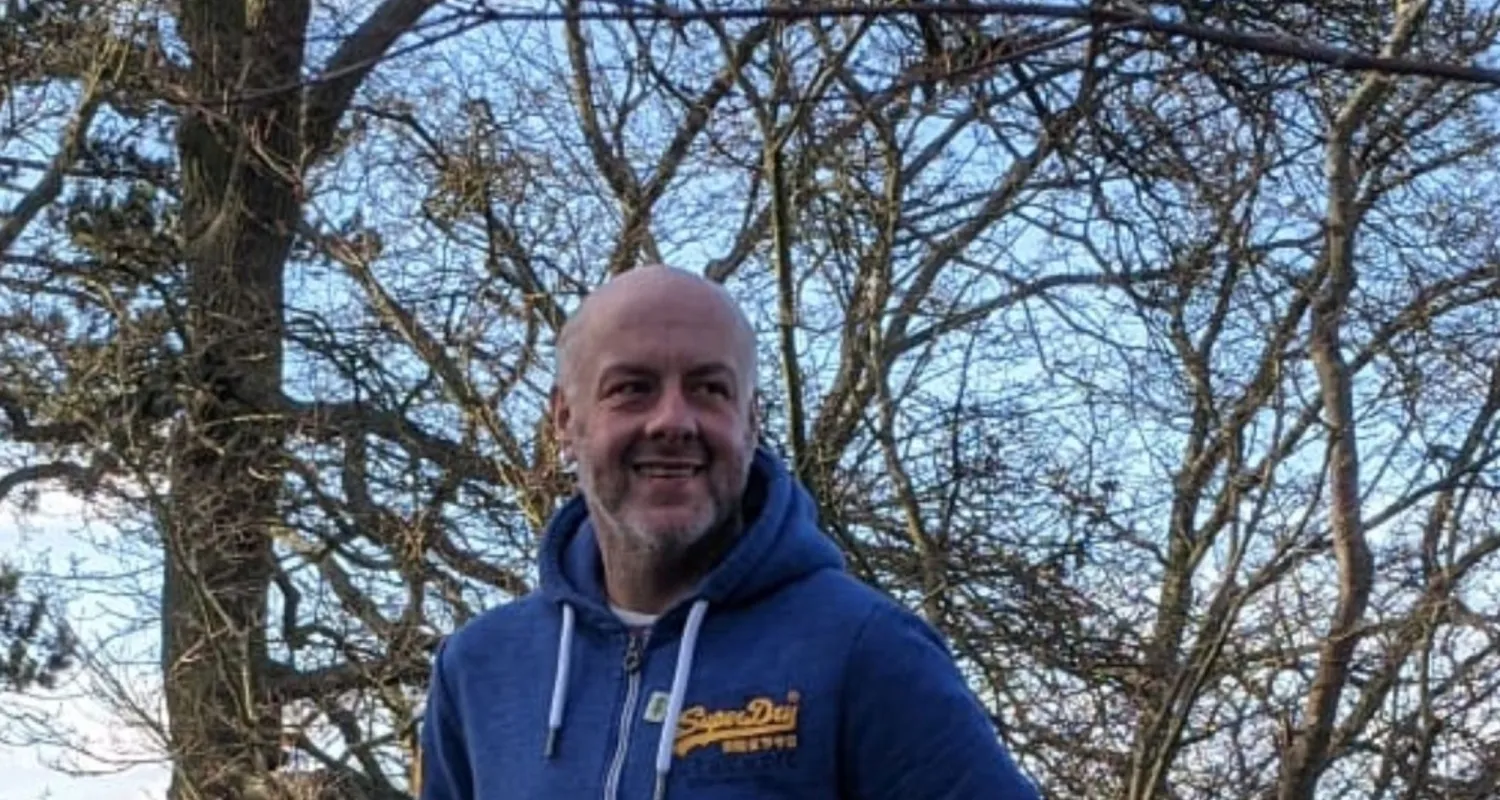 David smiling outside with trees in the background, wearing a zip-up hoodie