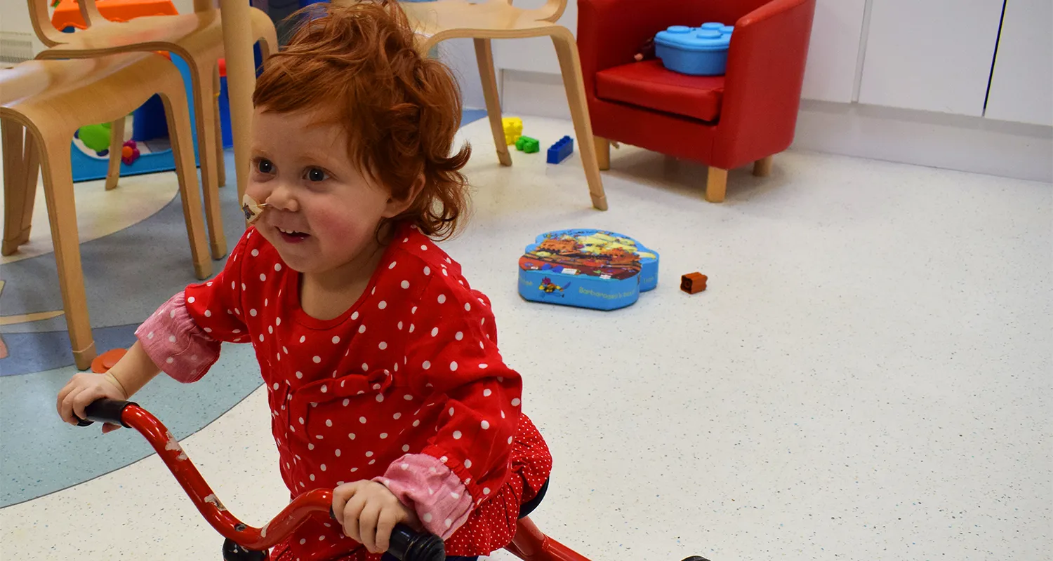Photo of Emily riding a tricycle in the playroom