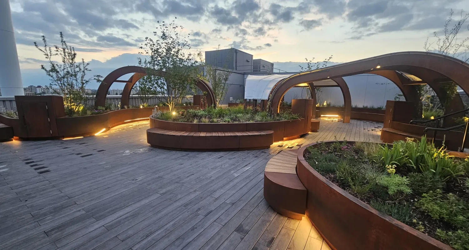 Modern rooftop garden with curved wooden benches and illuminated planters under a cloudy evening sky.