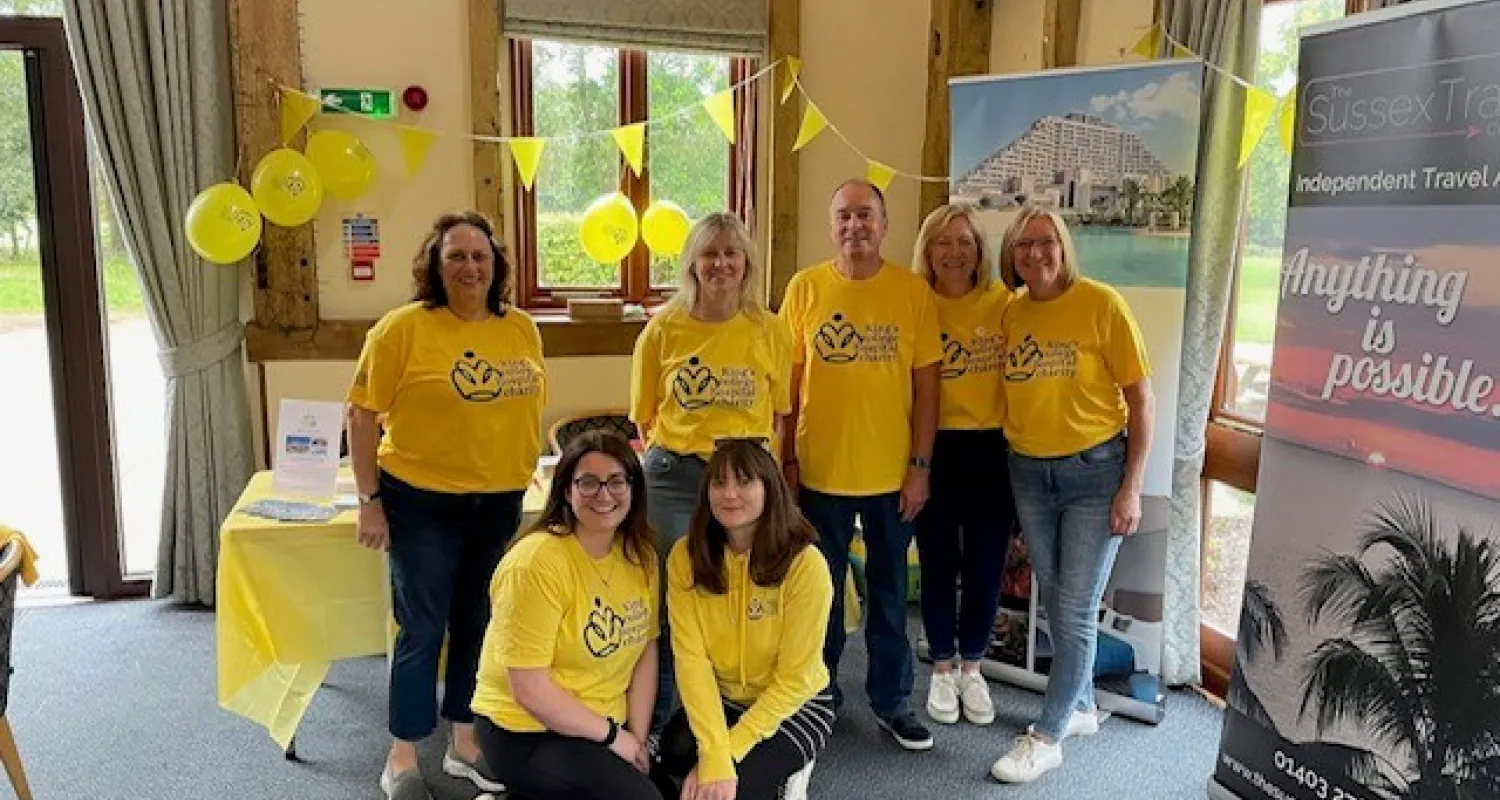 7 people in yellow t-shirts pose and smile at the camera in front of a table with yellow balloons