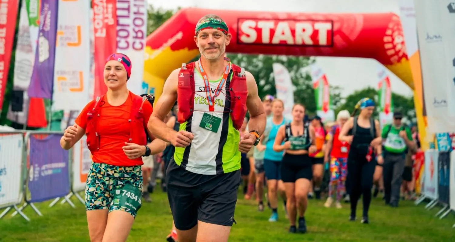 Two people jog towards the camera with flags and an event gantry behind them. 