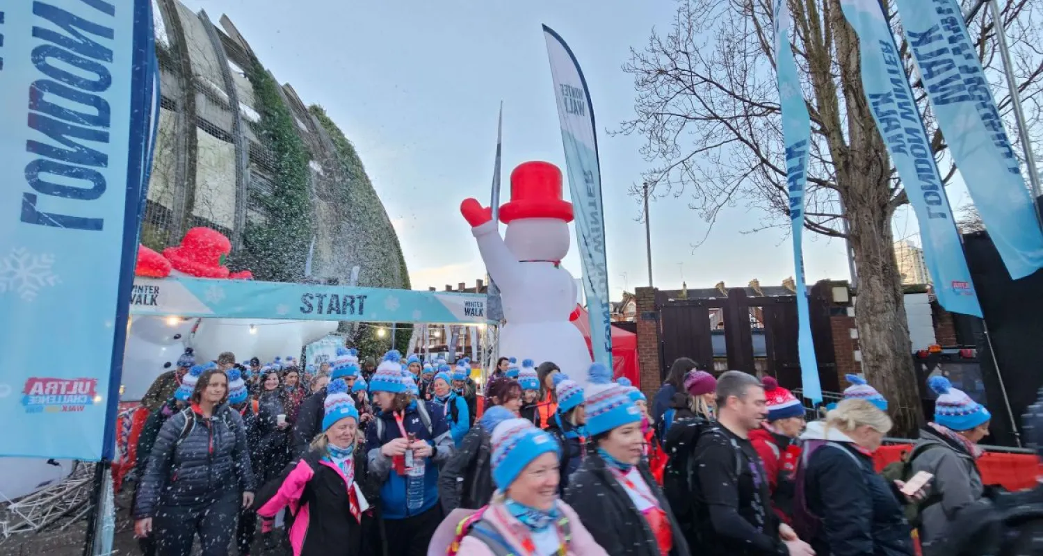 A group of people in winter clothes walk past the camera with event flags and a gantry behind them. 