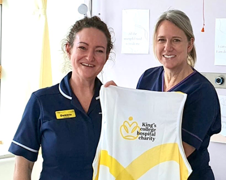 Two women (Debbie and Clare) in hospital scrubs holding up a King's College Hospital Charity running vest and smiling at the camera