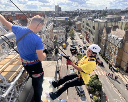 A woman leans of the roof attached to the abseil structure as she prepares to abseil with the instructor. The London skyline is behind her. 
