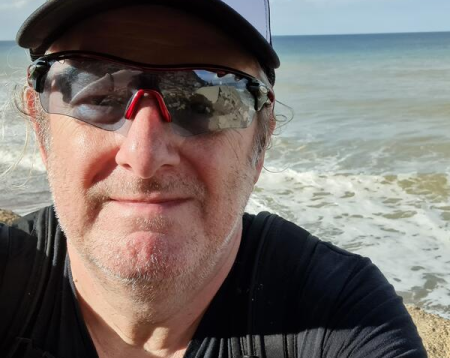 Photo of Ray with cap and sunglasses sitting on beach with sea behind