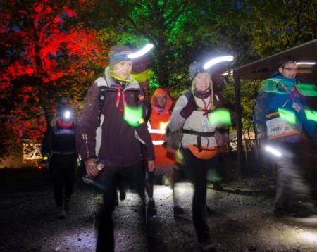 A group of people walk at night time, lit up with headtorches and other lights.