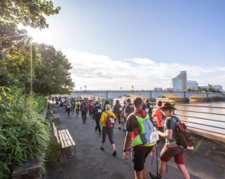 A group of walkers walk away from the camera with the Thames on their right.