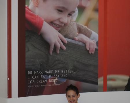 Reggie as a young child, standing in school uniform behind a large poster on the wall of him smiling, with a quote that reads: Dr Mark made me better. I can eat pizza and ice cream now. 