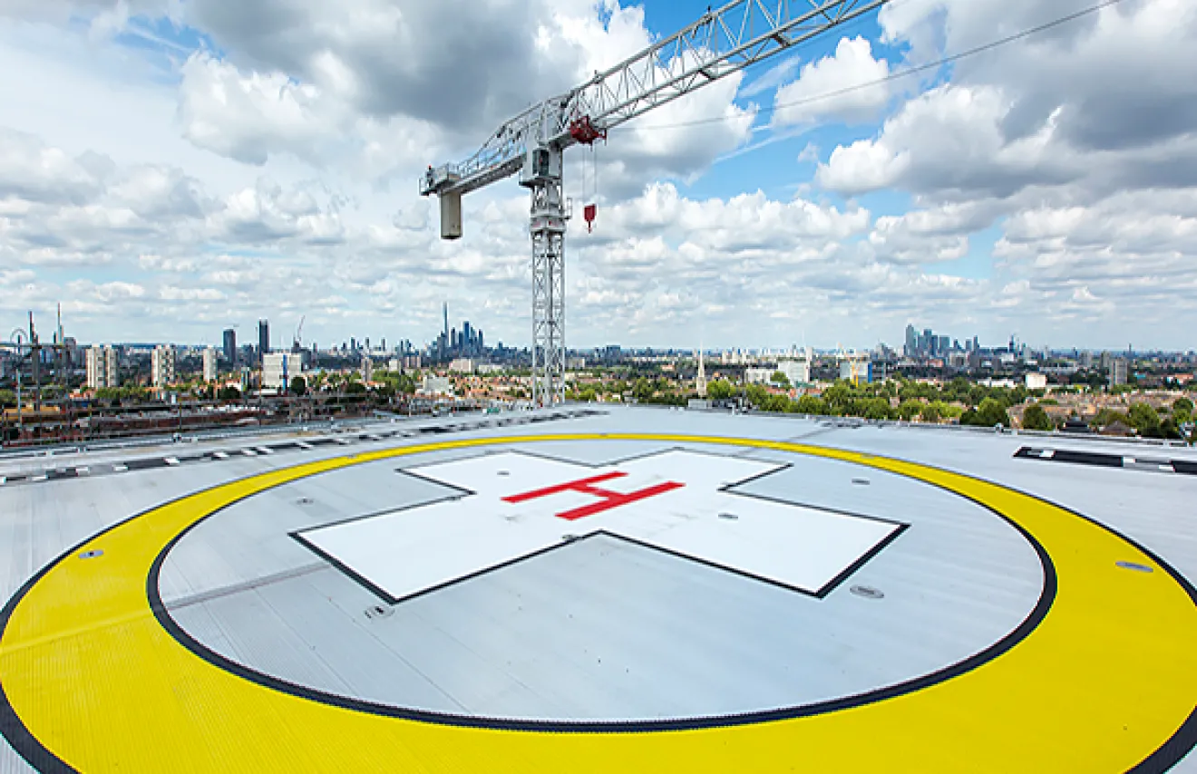 A birds-eye view of the helipad on a bright day with blue skies. There is a large yellow circle with a red cross in the middle to indicate where the landing strip is located for air ambulances. The London skyline is visible in the background.
