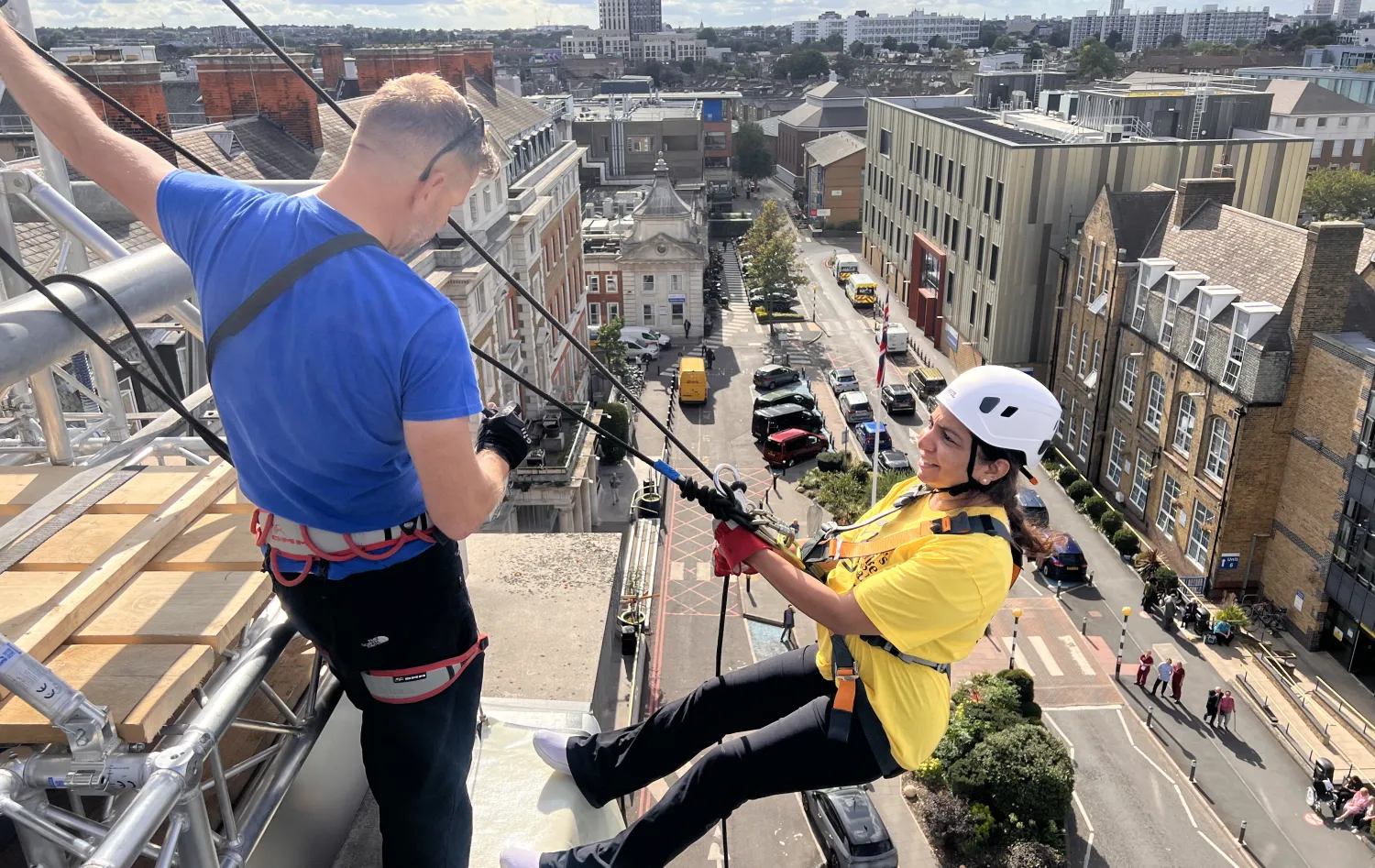 A woman leans of the roof attached to the abseil structure as she prepares to abseil with the instructor. The London skyline is behind her. 