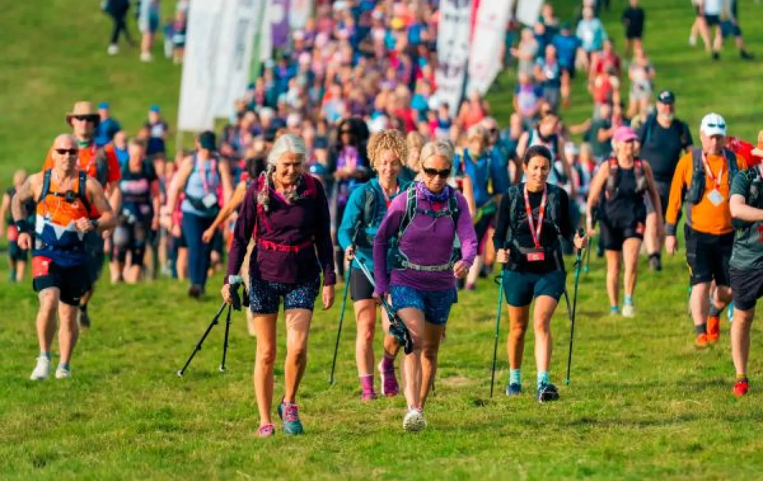 A large number of people walk towards the camera on grass, with flags in the background. 