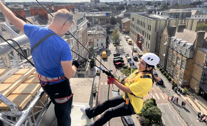 A woman leans of the roof attached to the abseil structure as she prepares to abseil with the instructor. The London skyline is behind her. 