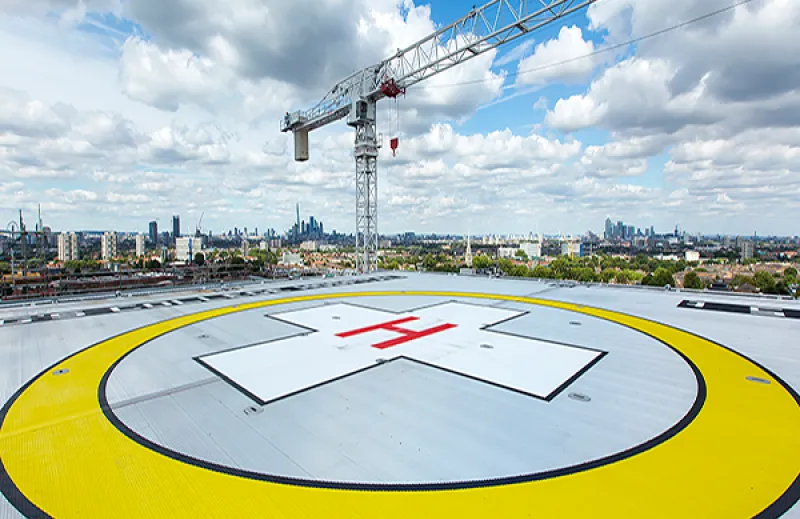 A birds-eye view of the helipad on a bright day with blue skies. There is a large yellow circle with a red cross in the middle to indicate where the landing strip is located for air ambulances. The London skyline is visible in the background.