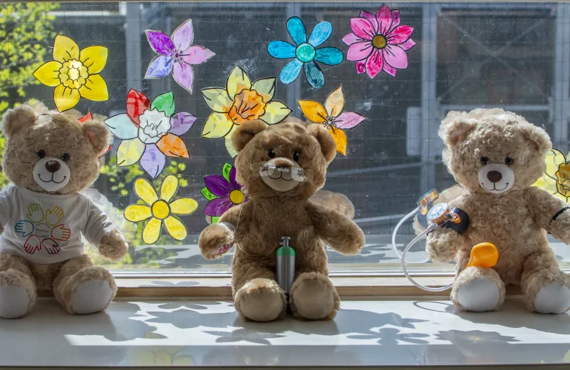 Three soft toy bears from the Teddy Bear Hospital pictured on a window sill, against a backdrop of colourful flower designs on the window glass 