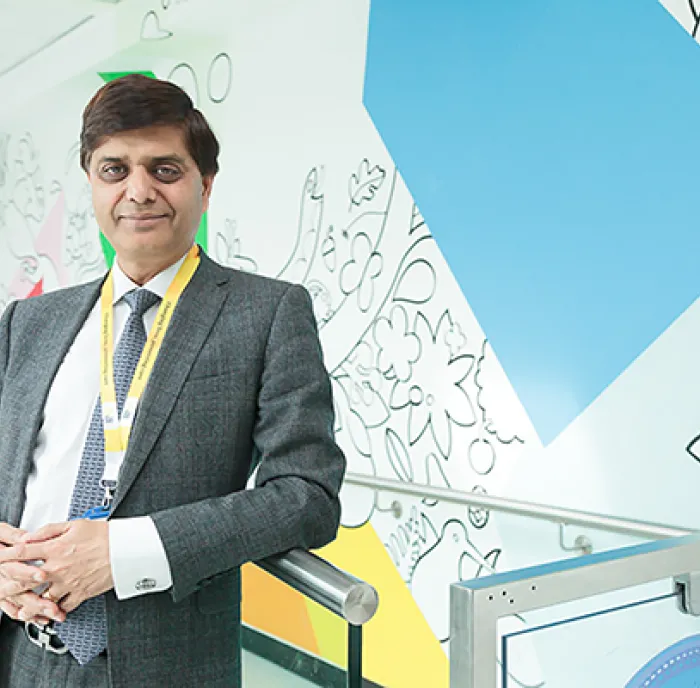 Professor Anil Dhawan, wearing a grey suit and blue tie, standing in a hospital corridor and smiling.