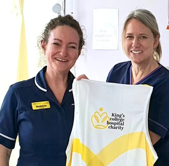Two women (Debbie and Clare) in hospital scrubs holding up a King's College Hospital Charity running vest and smiling at the camera