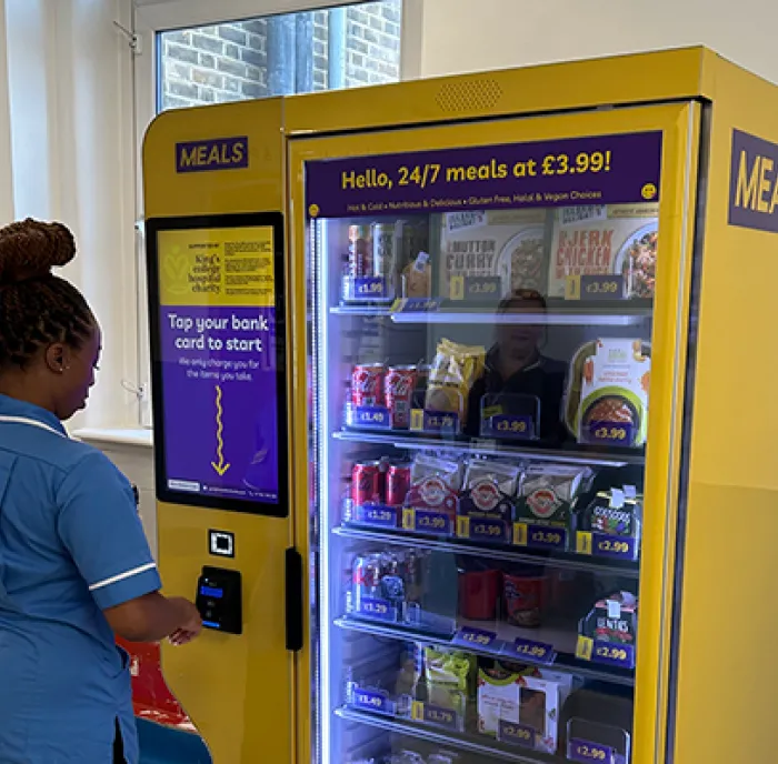 Female King's staff member in blue overalls using the smart fridge in the hospital corridor.