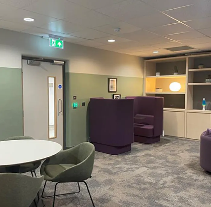 The brand-new Wellbeing Hub. The walls are a calming sage green and the carpet is a calming grey. There are twin chairs in a rich purple in the far left hand corner. In the near left corner there is a round, white table with three sage green soft chairs. There are two lava lamps in the back, as well as some more purple chairs to the right. There are calming pictures framed on the wall.