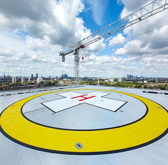 A birds-eye view of the helipad on a bright day with blue skies. There is a large yellow circle with a red cross in the middle to indicate where the landing strip is located for air ambulances. The London skyline is visible in the background.