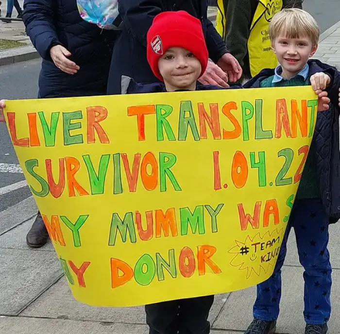 Picture of a child holding up a sign saying 'Liver Transplant Survivor 1.4.2022 - my mummy was my donor'