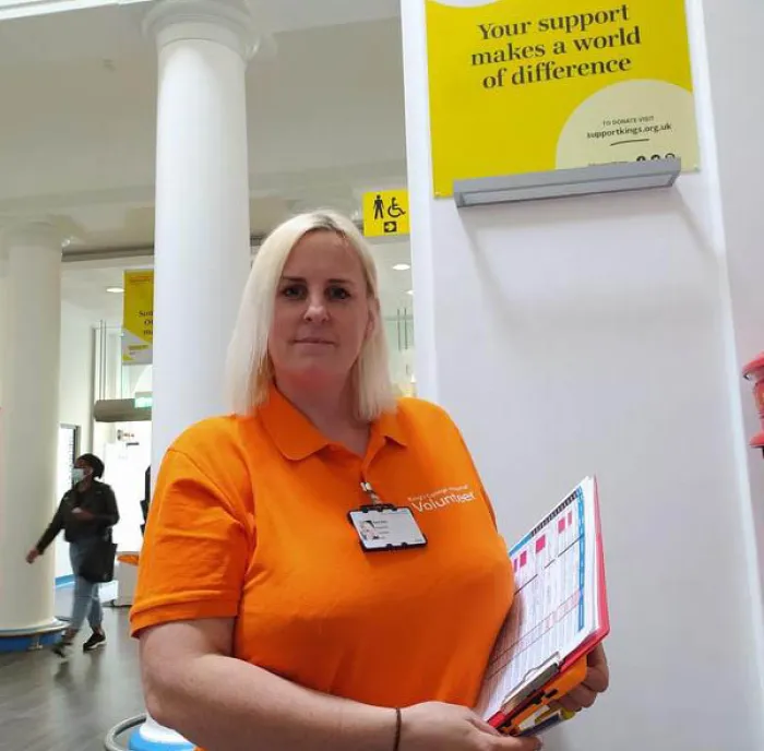 Photo of Rachel wearing orange King's Volunteer uniform standing holding clipboard in entrance to hospital