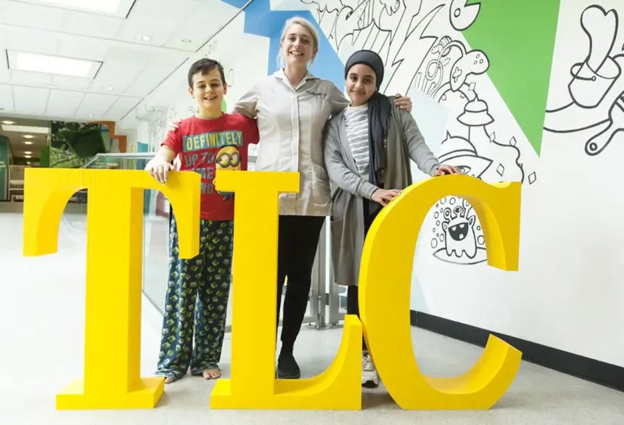 Picture of a nurse standing with her arms round two children, all standing behind one-metre high letters spelling T L C