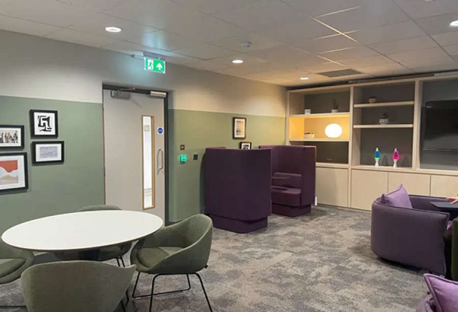 The brand-new Wellbeing Hub. The walls are a calming sage green and the carpet is a calming grey. There are twin chairs in a rich purple in the far left hand corner. In the near left corner there is a round, white table with three sage green soft chairs. There are two lava lamps in the back, as well as some more purple chairs to the right. There are calming pictures framed on the wall.