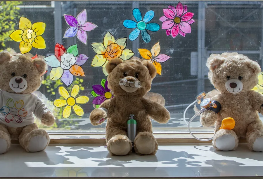Three soft toy bears from the Teddy Bear Hospital pictured on a window sill, against a backdrop of colourful flower designs on the window glass 