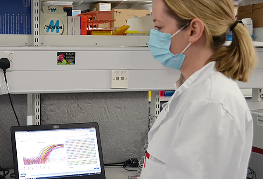 Picture of a woman in lab coat and mask pointing at a colourful graph on a computer screen