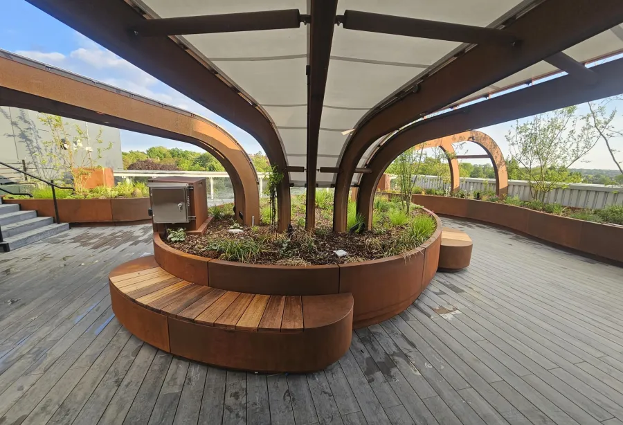 Curved wooden benches surround a central planter with metal arches and a canopy overhead on a rooftop deck.