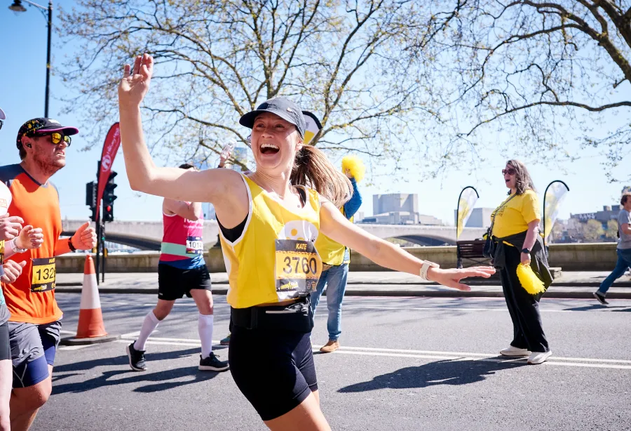 A lady in a hat and yellow vest is running and smiling broadly at someone off camera
