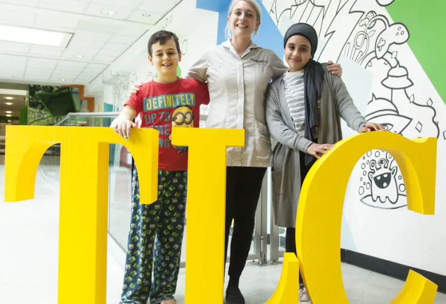 Picture of a member of the liver care team standing with arm round two young patients standing in front of giant letters T L and C