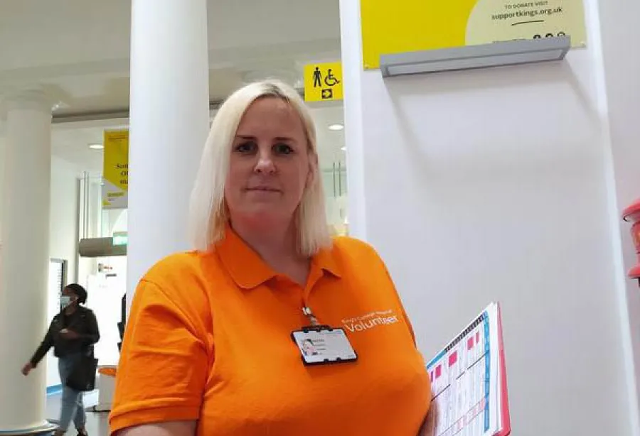 Photo of Rachel wearing orange King's Volunteer uniform standing holding clipboard in entrance to hospital